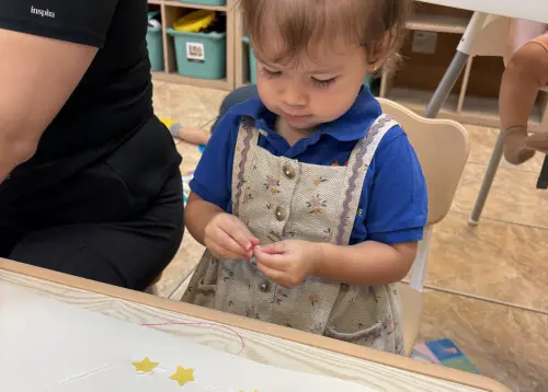 A young toddler girl focused on a glue and paper star craft activity at a Miami Springs daycare table.